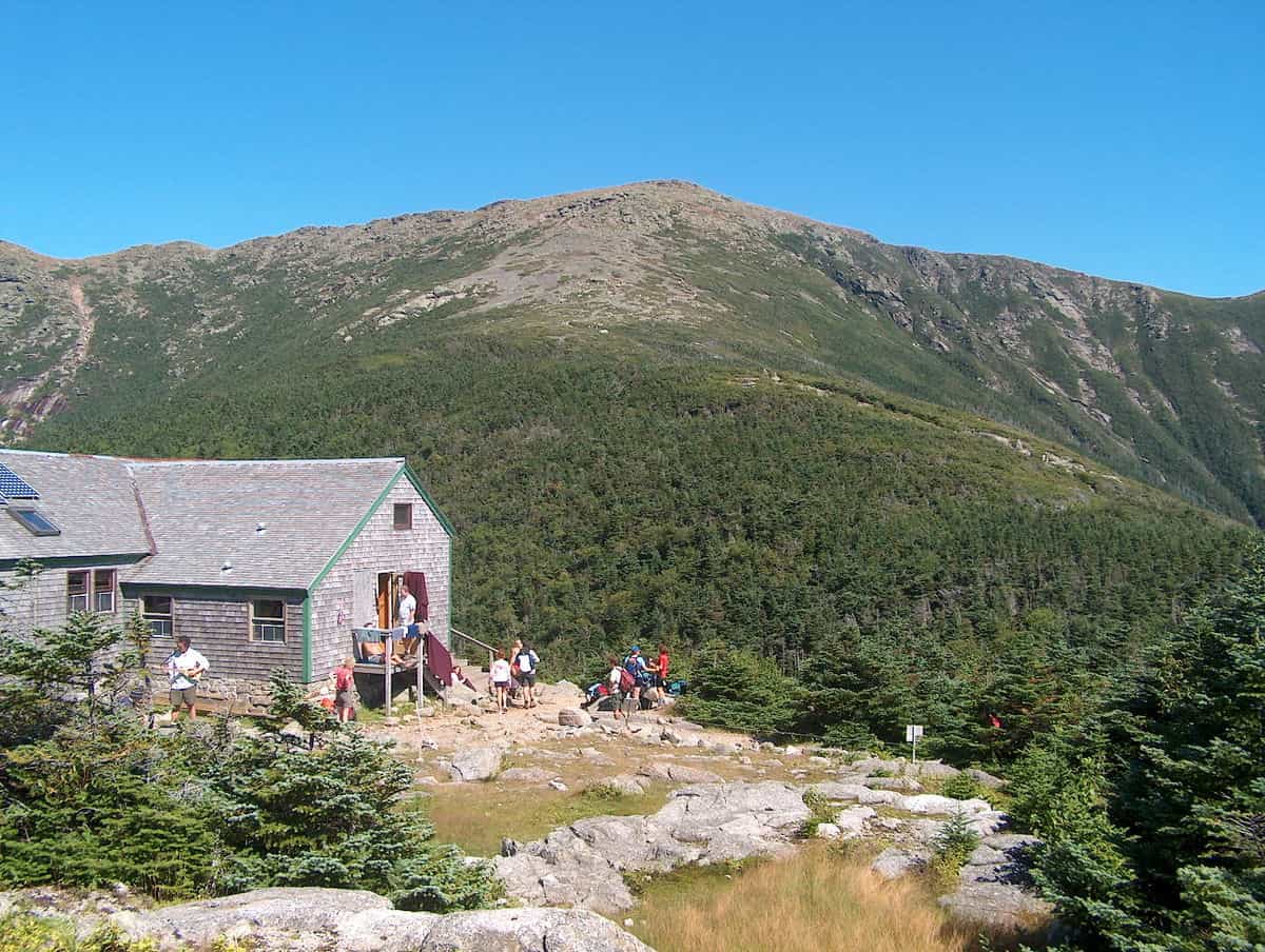 Hikers resting outside Greenleaf Hut below a large, tree-covered mountain peak.