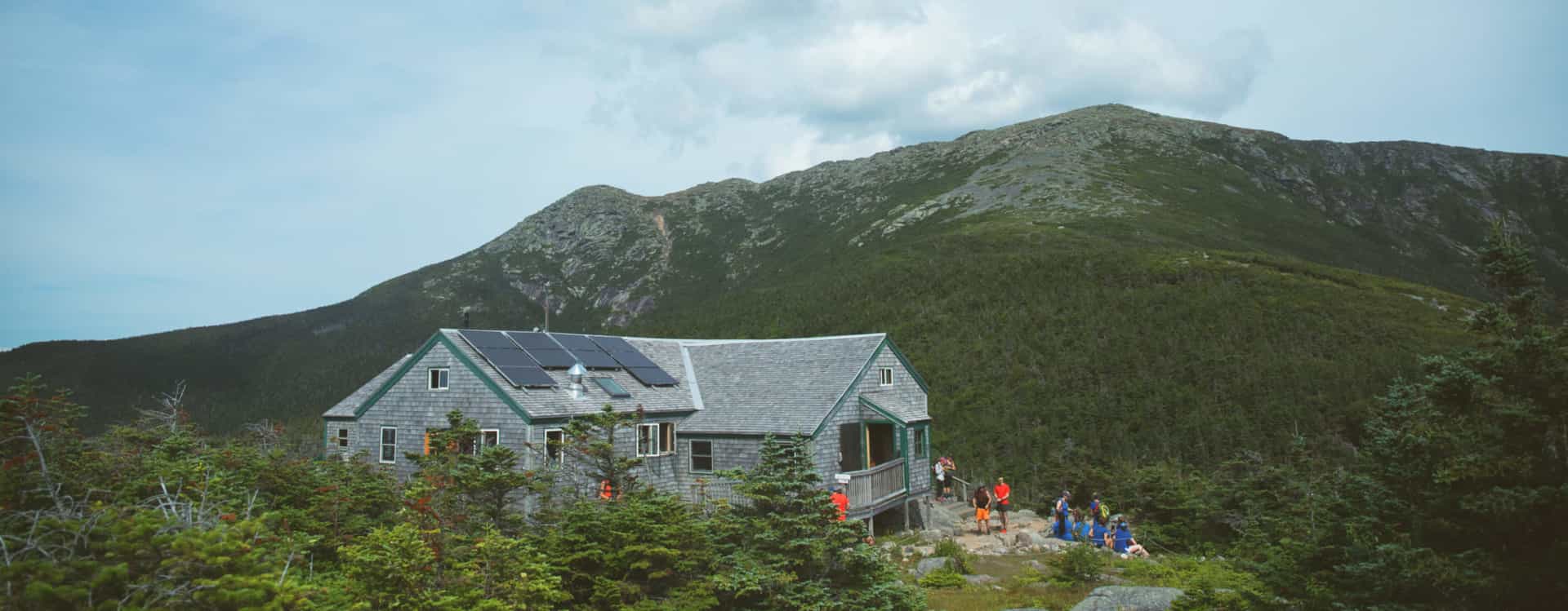 Mountain hut with solar panels nestled in dense forest below a large, rocky peak.