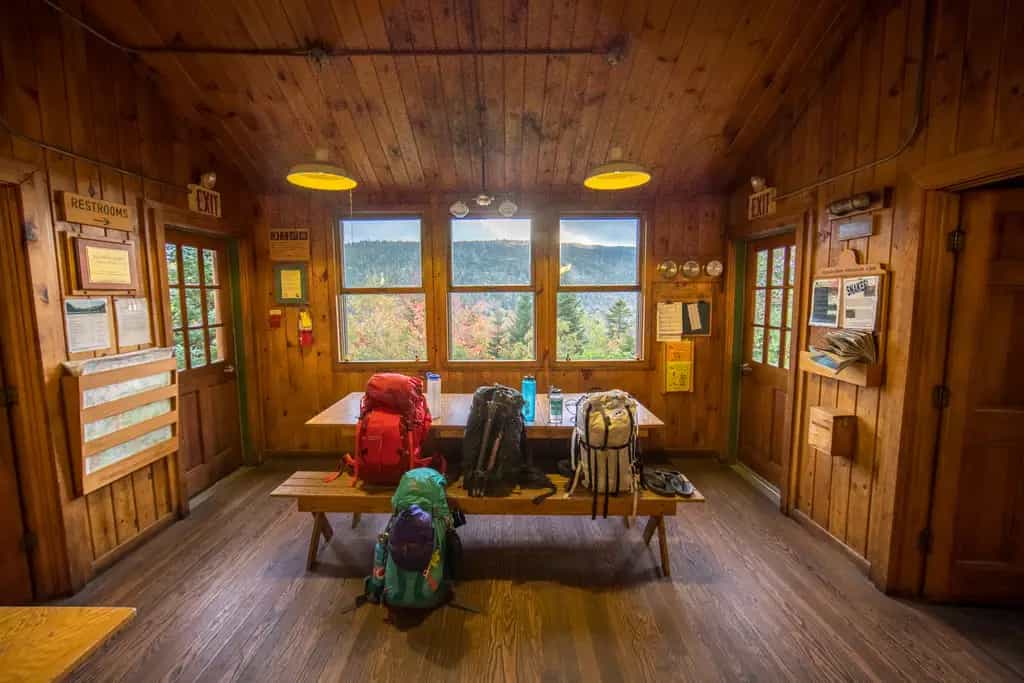 Backpacks inside Zealand Falls Hut with view of autumn forest mountains.