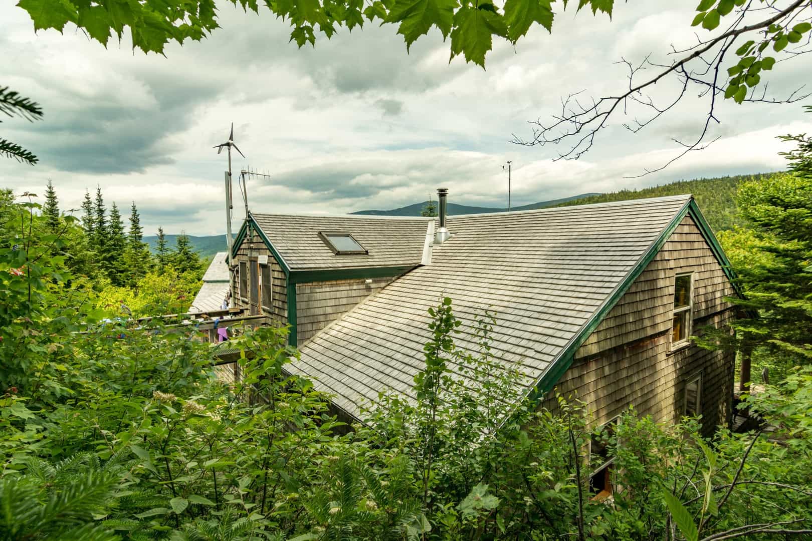 Zealand Falls Hut with cedar shingles surrounded by dense green foliage and distant mountains.