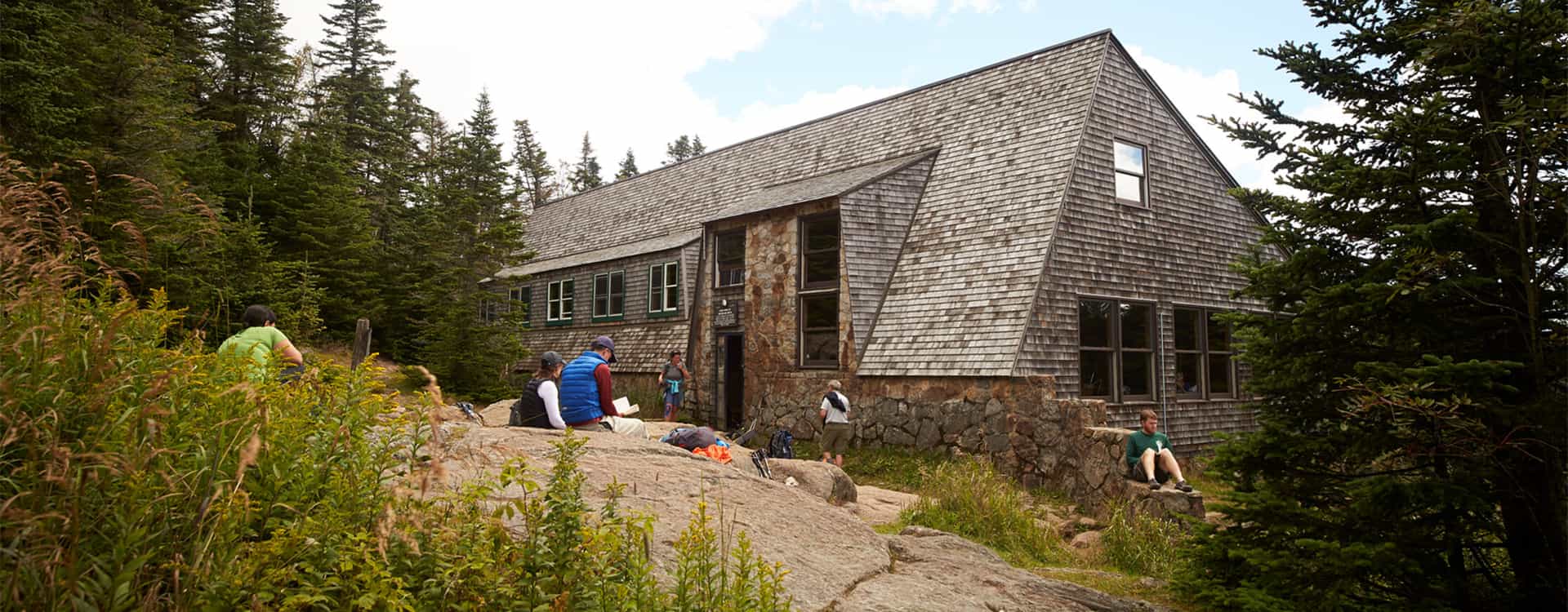Hikers resting outside Mizpah Spring Hut surrounded by dense evergreen forest and rocks.