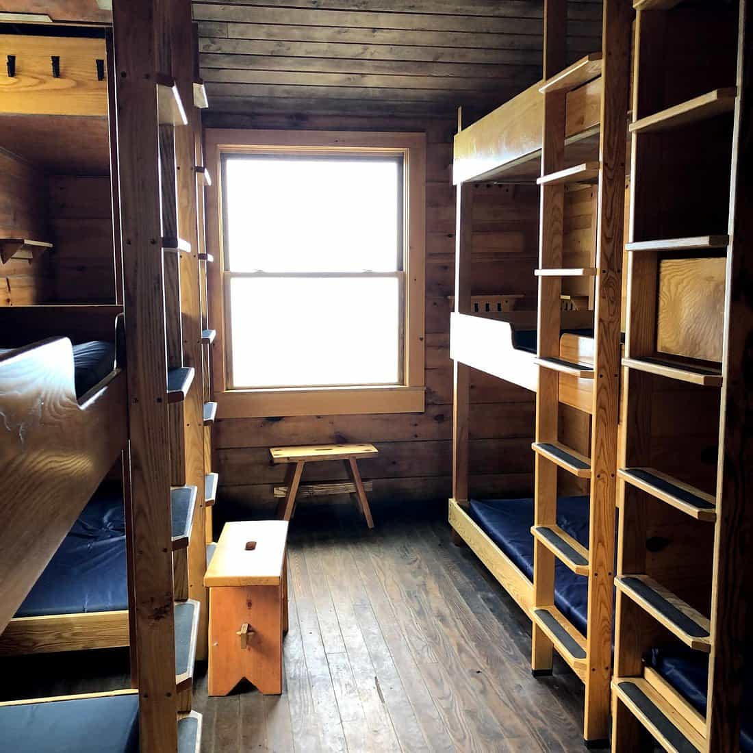 Interior of Lakes of the Clouds Hut showing wooden bunk beds, stools, and a bright window.