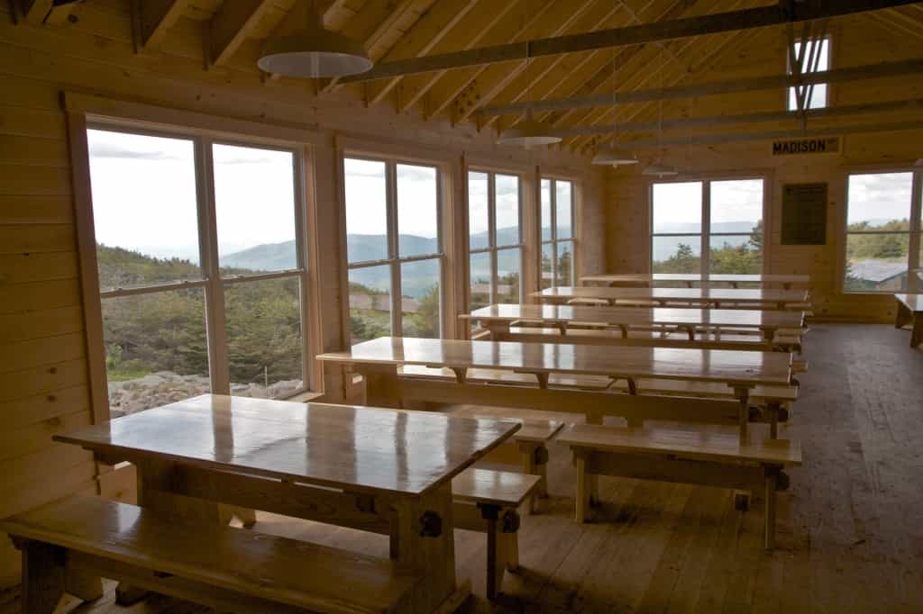 Wooden dining hall with long tables and benches inside Madison Spring Hut overlooking mountains.