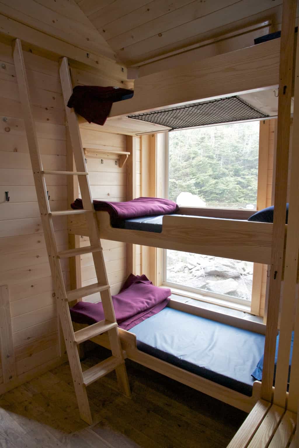Wooden bunk beds with purple blankets inside a cabin with a view of a forest.