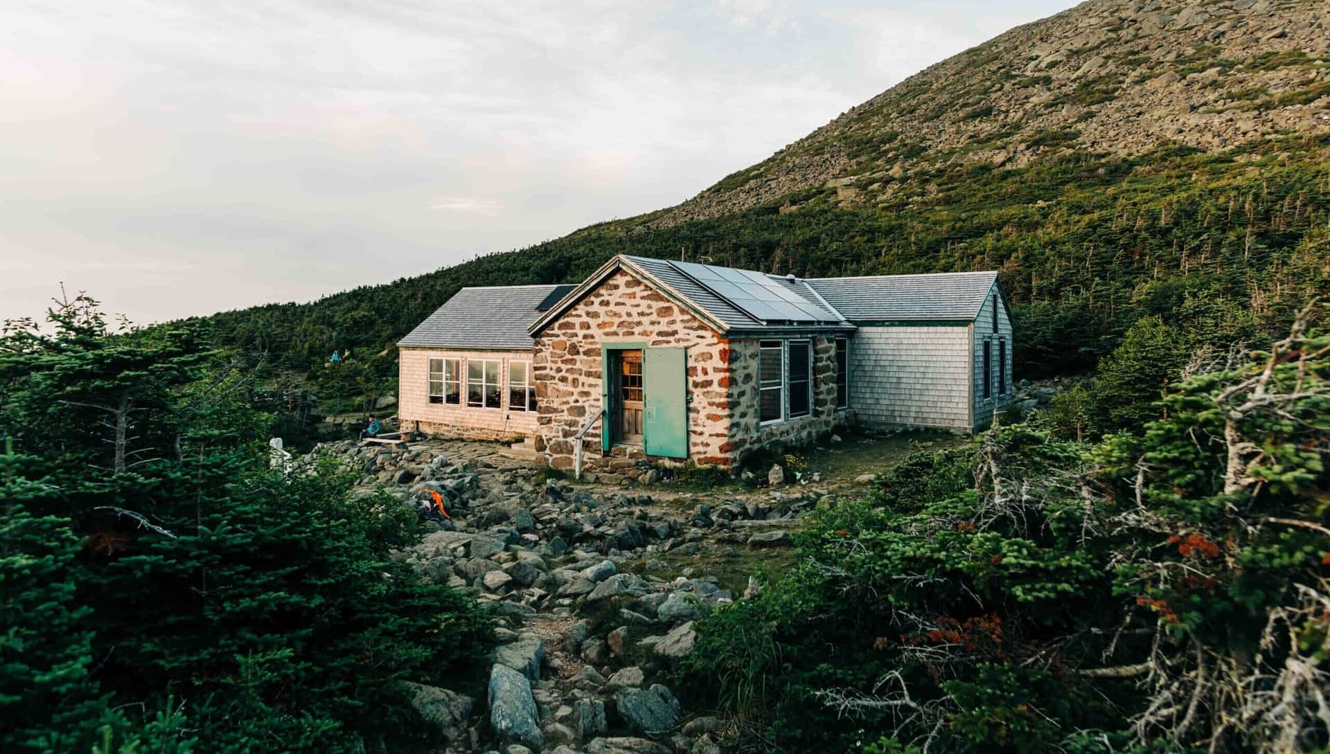 Stone and shingled mountain hut with solar panels nestled among dense green trees and boulders.