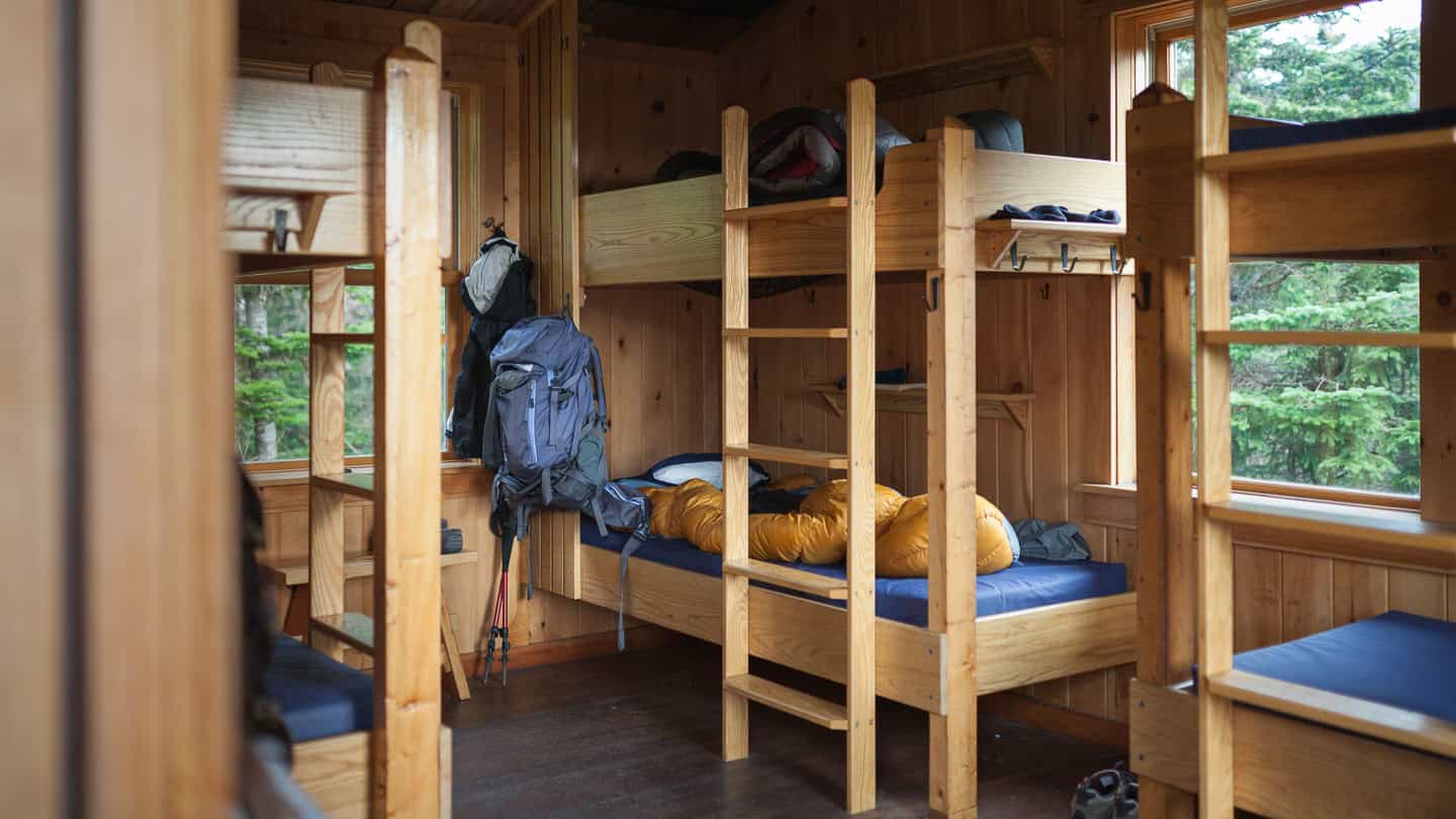 Wooden bunk beds inside a rustic hut with backpacks and sleeping bags, forest view.