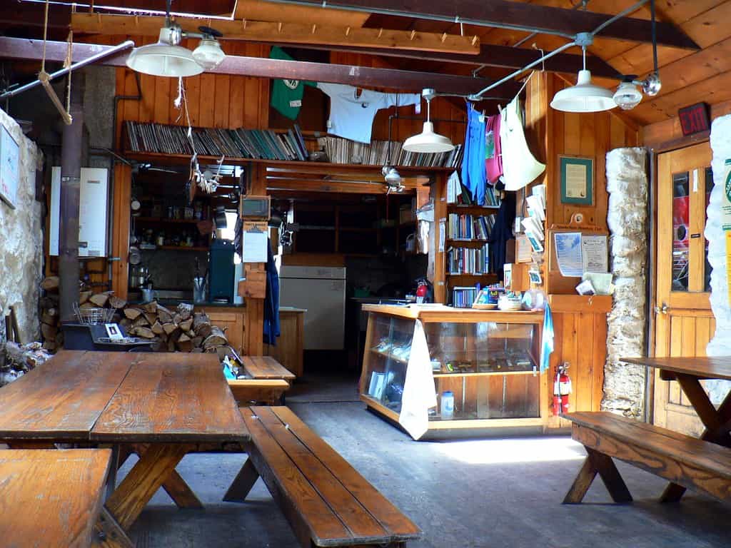 Interior of a rustic wooden mountain hut with picnic tables, firewood, and clothes drying overhead.