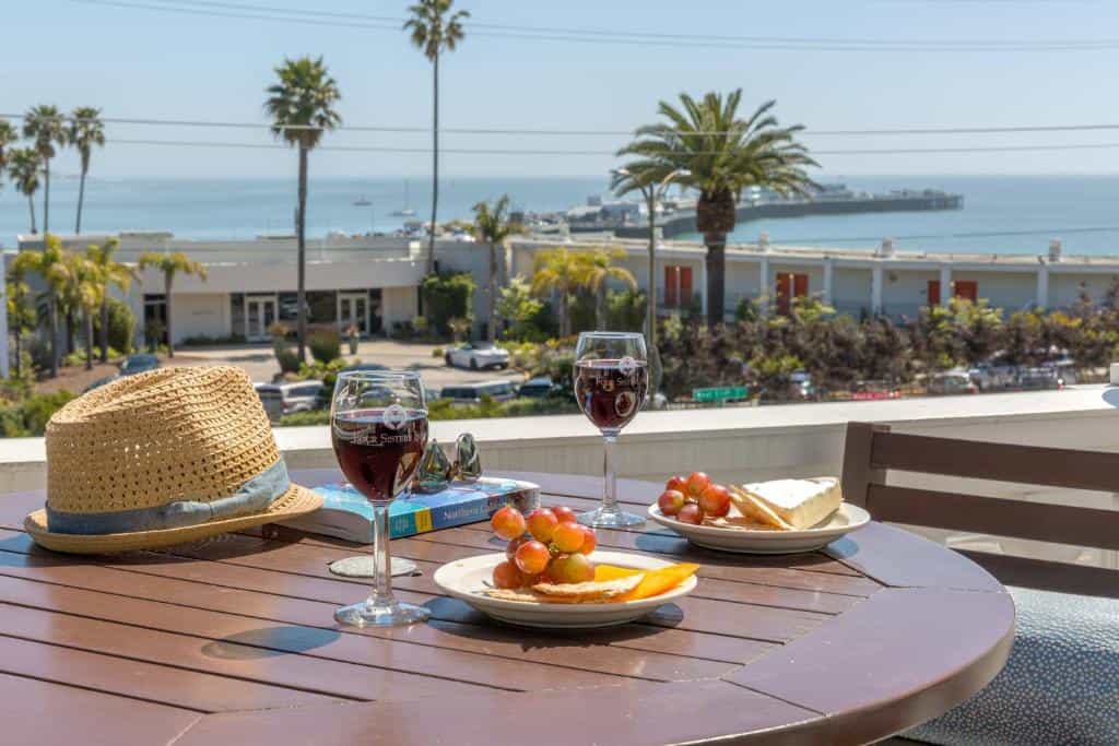 Wine, grapes, and cheese on a table overlooking a coastal pier and ocean.