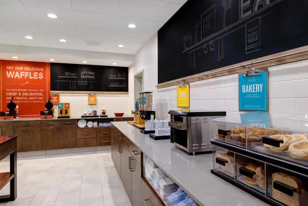 Hotel breakfast buffet station with waffle makers, cereal dispensers, and assorted baked goods.