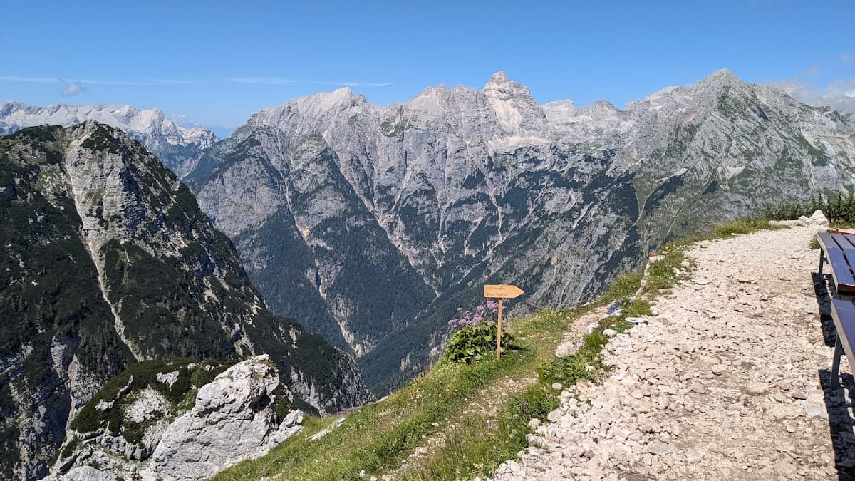 Rocky mountain trail overlook with wooden signpost and steep valley view under blue sky.
