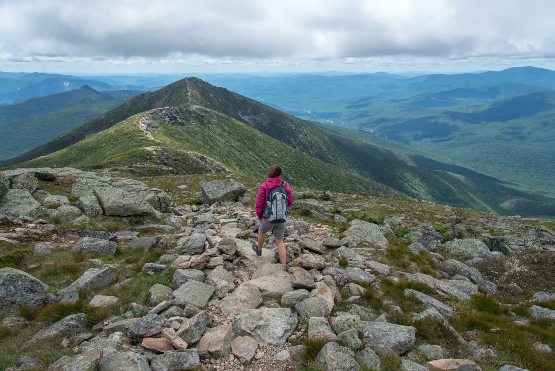Hiker on rocky trail ascending Mount Lafayette with expansive green mountain ranges below.