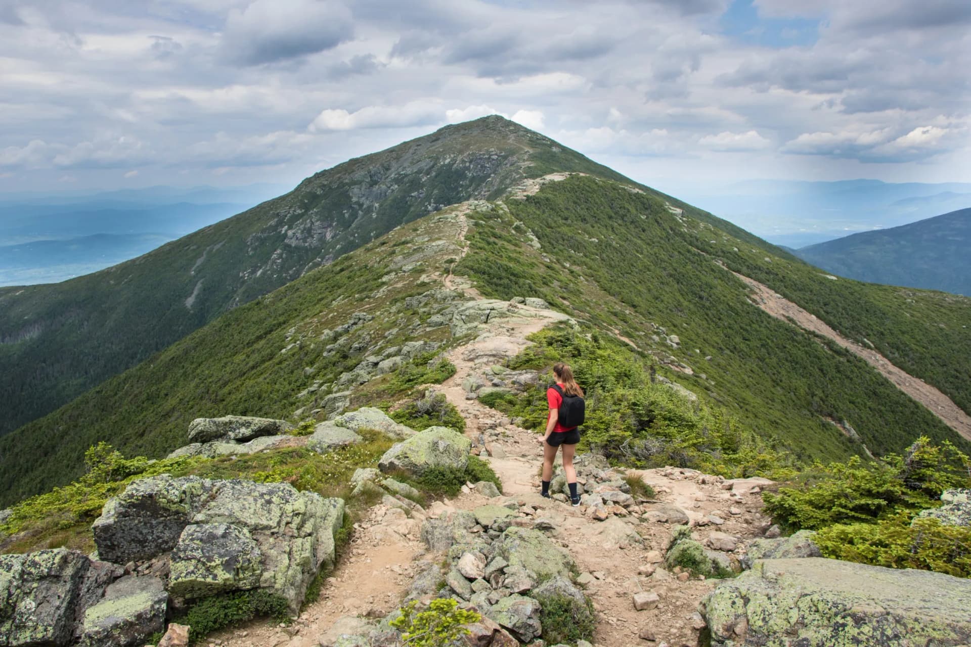 Hiker walking on rocky trail along a green, steep mountain ridge under cloudy sky.
