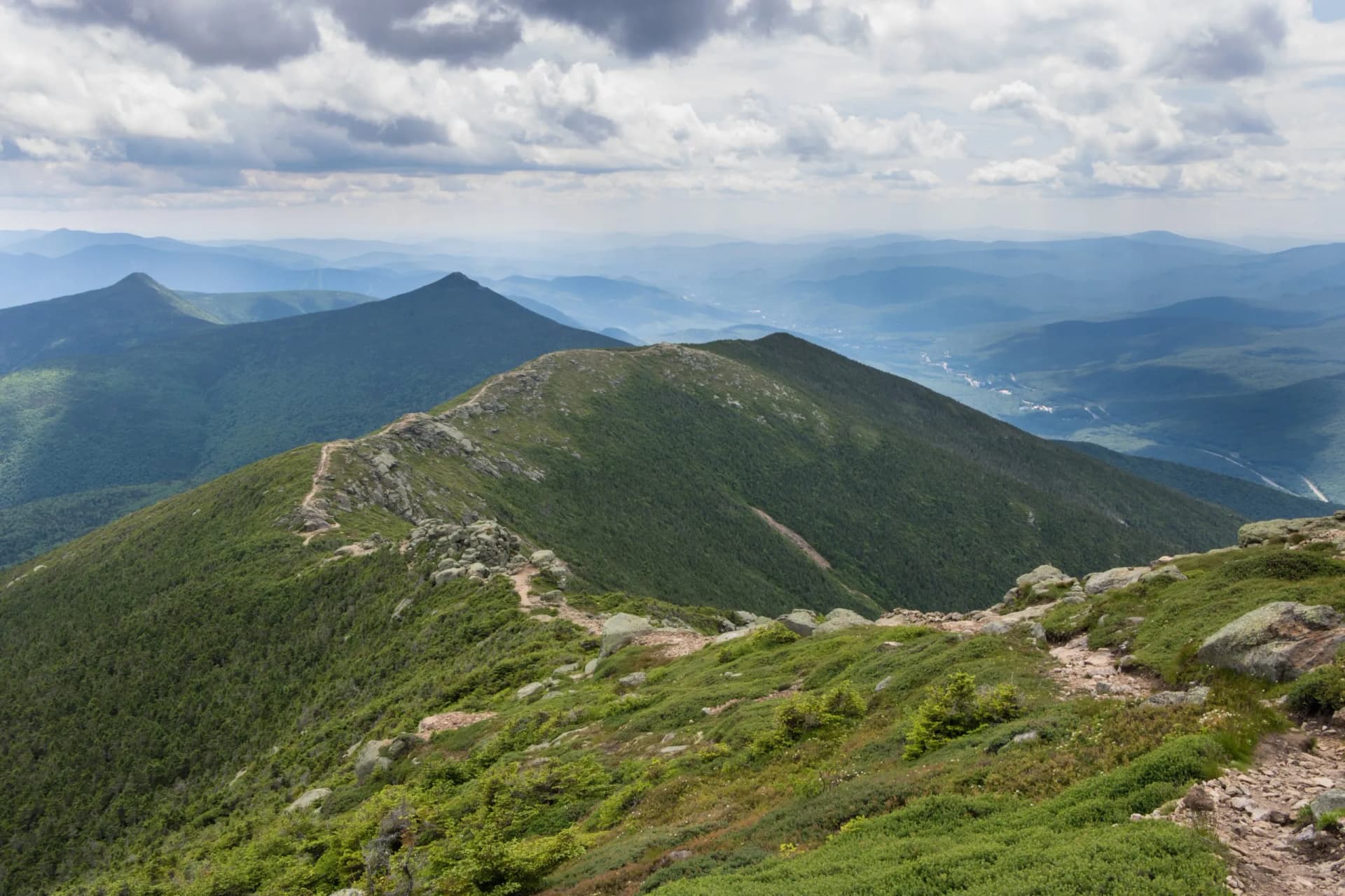 Hiking trail along a rocky, green ridgeline with layered blue mountains in New Hampshire.
