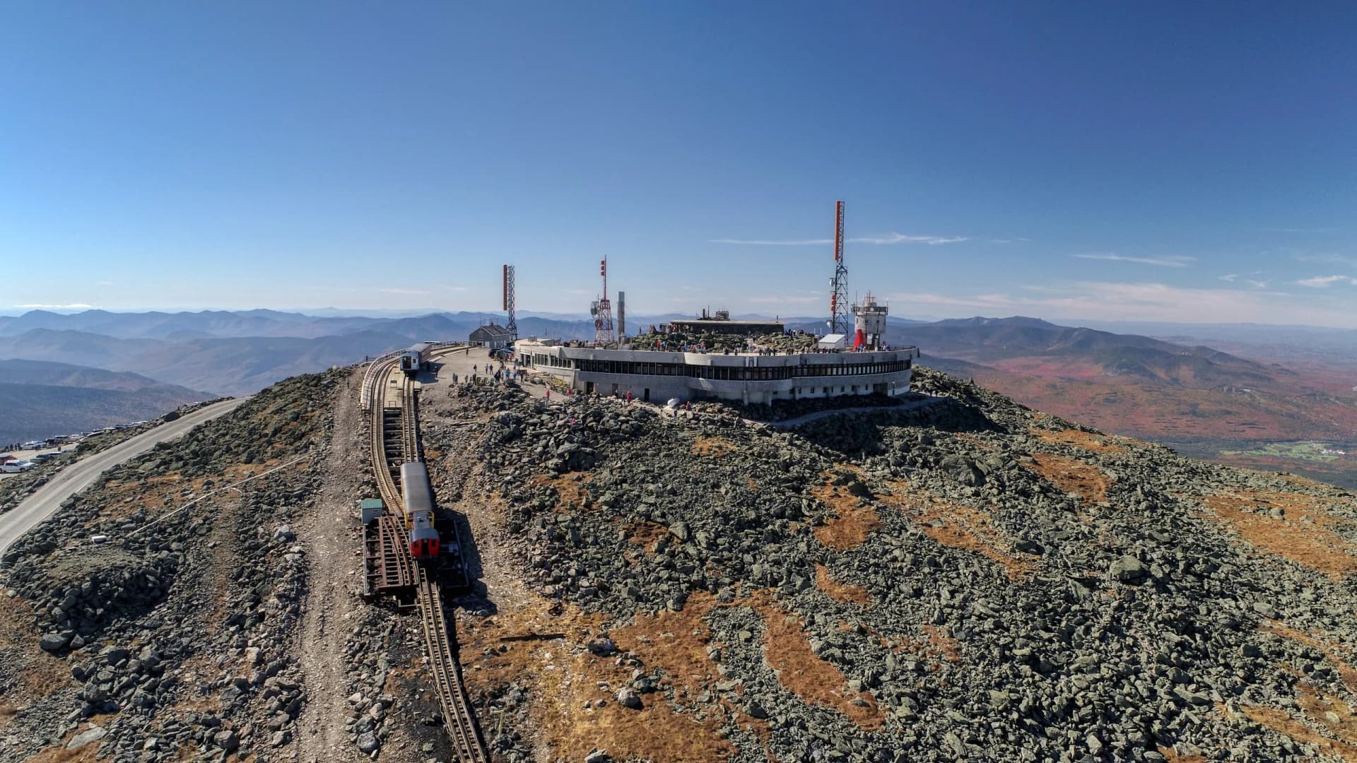Cog Railway train at Mount Washington summit building with rocky terrain and distant blue mountains.