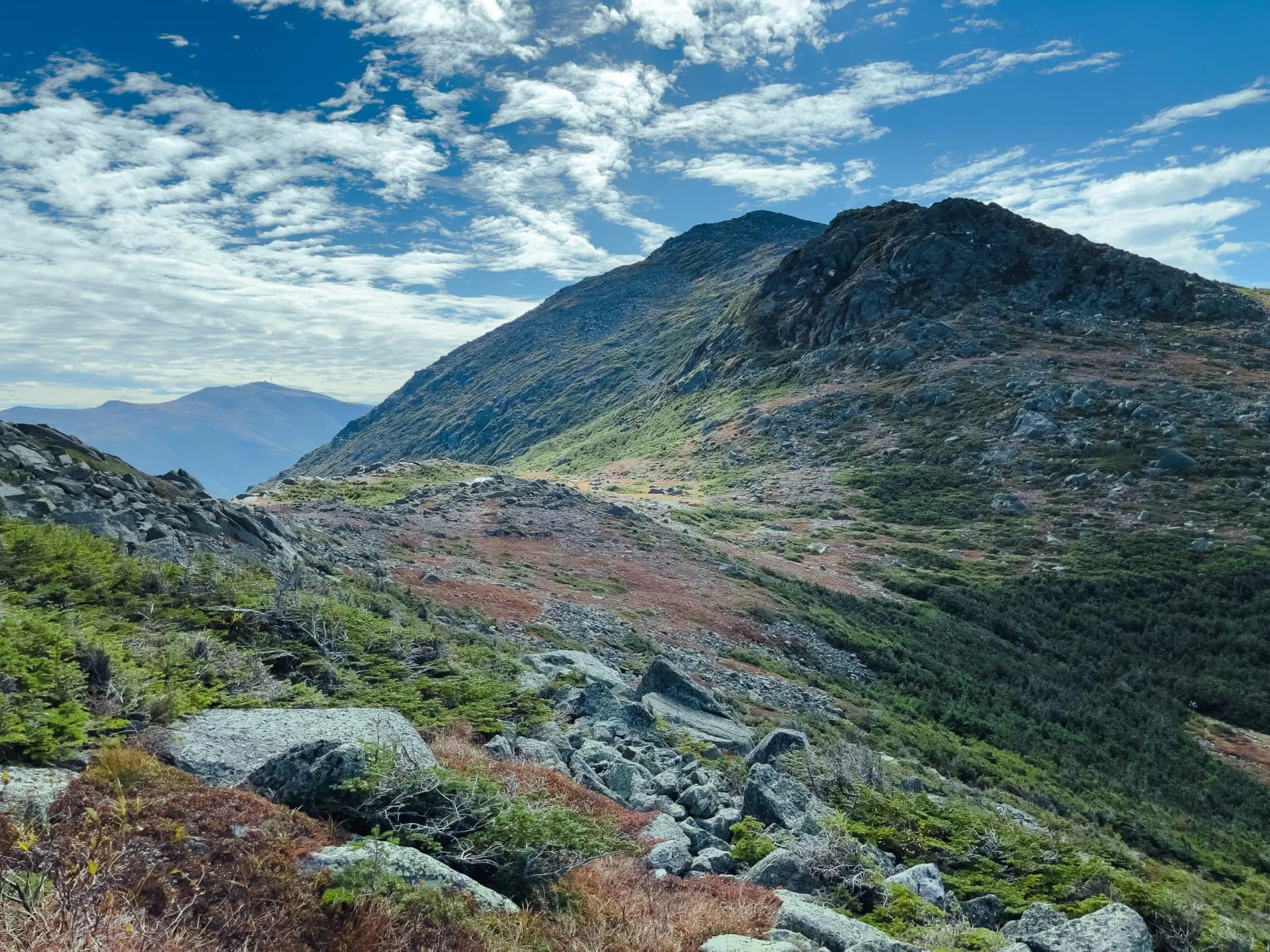 Rocky alpine terrain with sparse vegetation under a bright blue sky with white clouds, showing Mt. Adams, Mt. Madison, and...