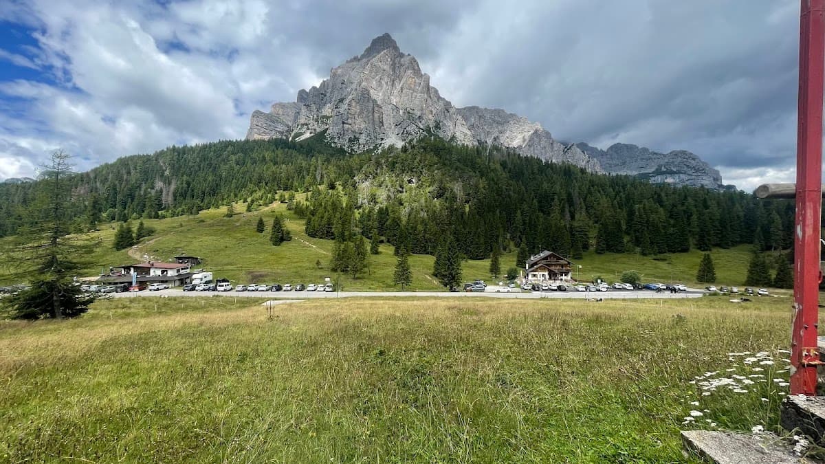 Alpine meadow with buildings and parking lot below a dramatic, rocky mountain peak.