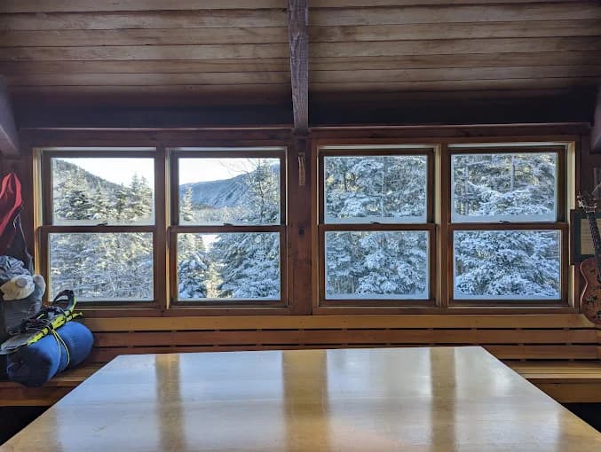 Wooden cabin interior with large windows looking out at snowy pine trees and mountains