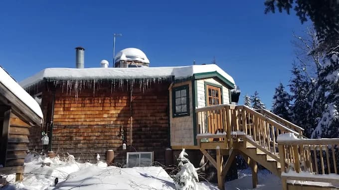 Wooden cabin with snow-covered roof and outdoor wooden stairs in winter forest