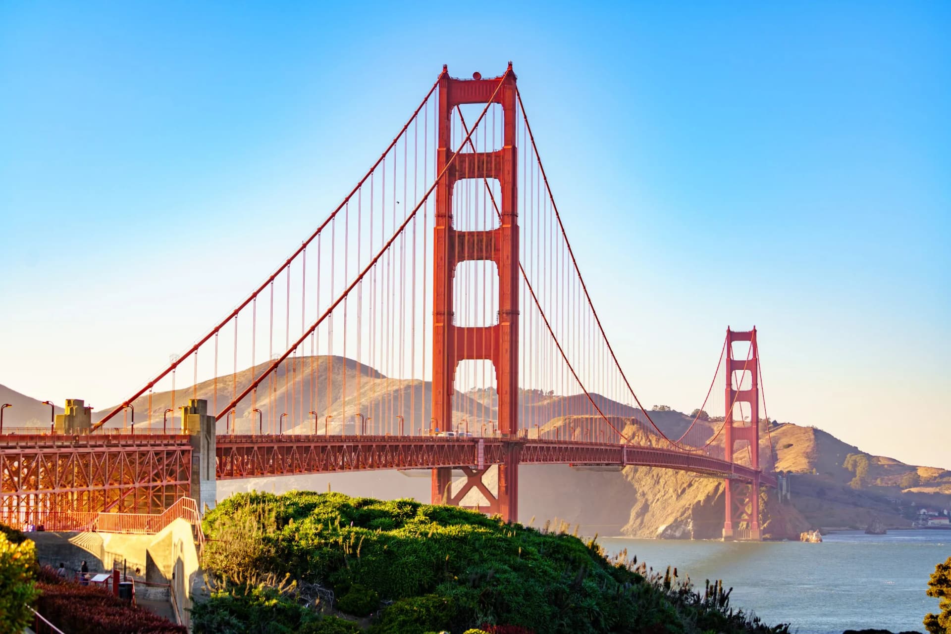Golden Gate Bridge spanning water with hills in the background under a clear blue sky.
