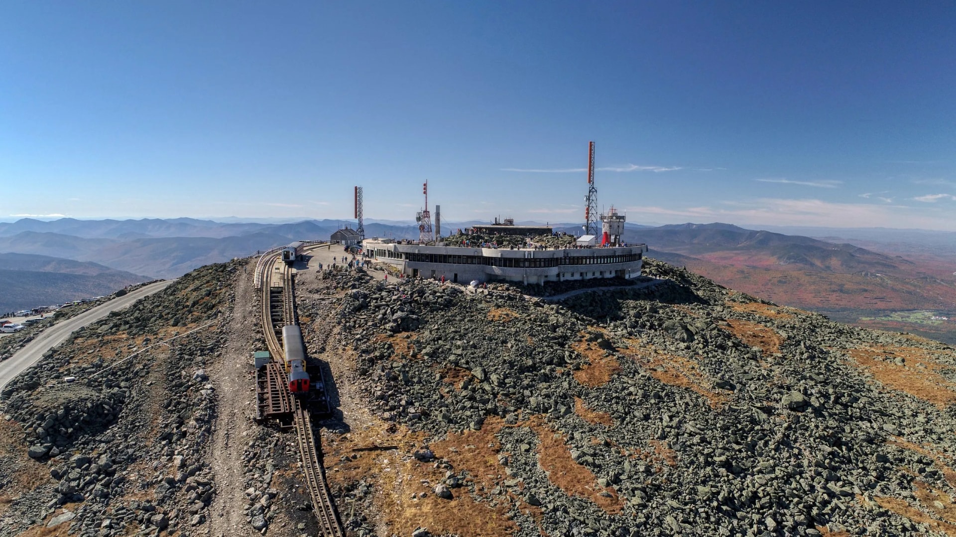 Cog Railway train approaching summit building on rocky Mount Washington with distant blue mountains.