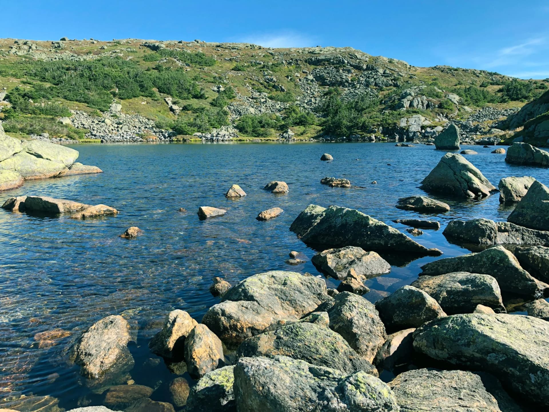 Clear water of Lake of the Clouds with rocks in foreground and scrubby mountains under blue sky at Mount Monro.