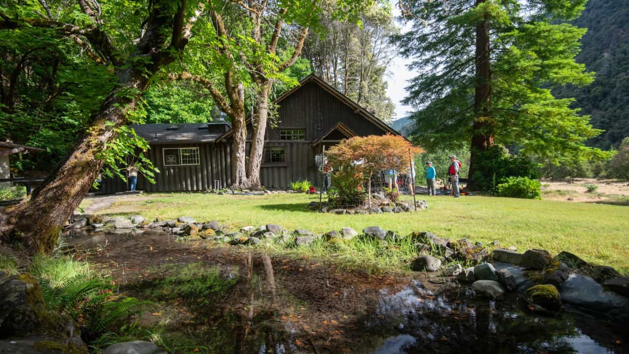 Rustic wooden lodge surrounded by lush green forest and trees near a small pond.