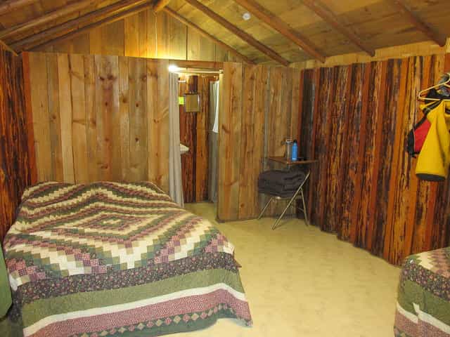 Rustic cabin interior with wood paneling, two beds with patterned quilts, and hanging outdoor gear.
