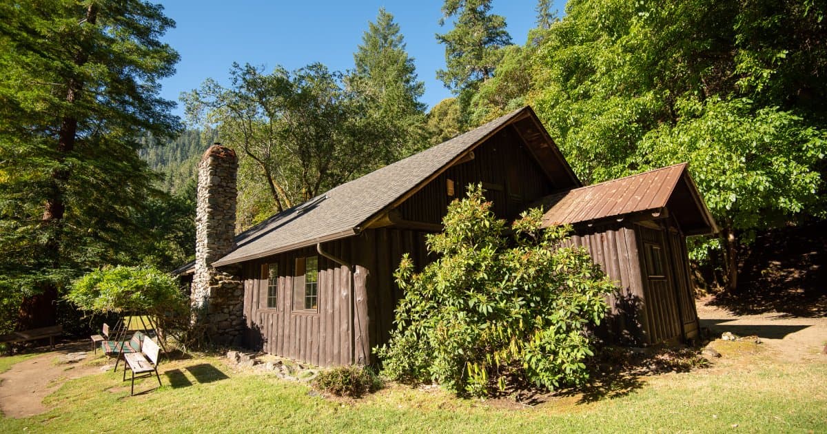 Rustic wooden lodge with stone chimney surrounded by dense green forest and tall trees.
