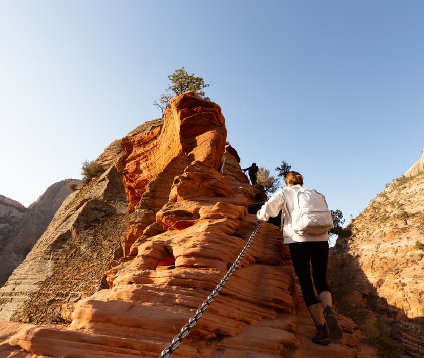 Hiker climbing steep red sandstone rock formation holding onto a chain in Zion National Park